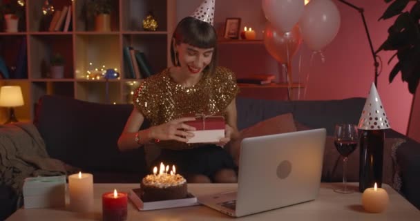 Young cheerful woman looking to laptop screen and talking while celebrating birthday online. Happy female person in paper hat opening present box and rejoicing while having video call. — Stock Video