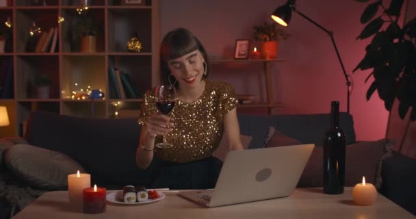 Dizüstü bilgisayar ekranına bakarken camdan şarap içen güzel bir genç kadın görüntüsü. Evde internetten iletişim kuran şık giyinmiş neşeli bir kadın.. — Stok video