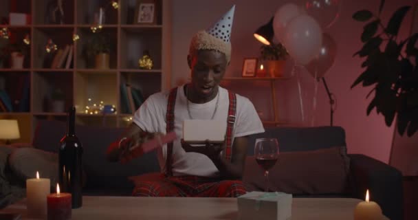 Man in birthday hat opening present and talking while looking to camera. Millennial positive guy with dyed hair saying wow and rejoicing while sitting at home. Concept of online party. — Stock Video