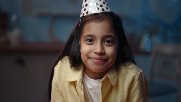 Widok na zboże małej wesołej dziewczyny uśmiecha się i patrzy do kamery. Cute dziecko w kapeluszu urodzinowym raduje się podczas pozowania. Niewyraźne tło. — Wideo stockowe