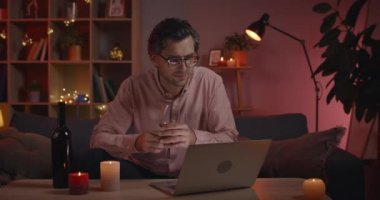 Laptop ekranına bakarken şarap kadehinden su içen yakışıklı bir erkek. Kanepede otururken gülümseyen ve sohbet eden gözlüklü olumlu bir adam. Çevrimiçi iletişim kavramı.