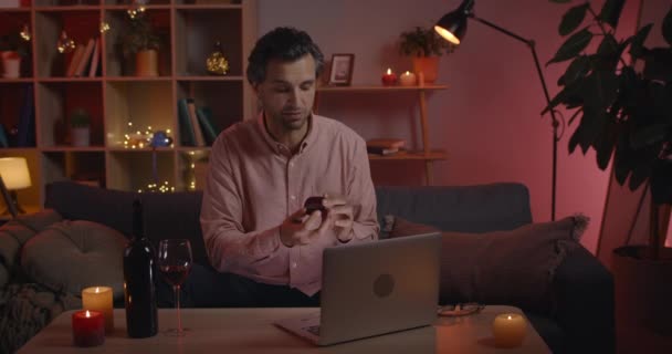 Προβολή των καλλιεργειών όμορφος καυκάσιος άνθρωπος κάνει online πρόταση γάμου, ενώ κάθεται στον καναπέ στο σπίτι. Guy στα 40 χαρά, ενώ κρατώντας το δαχτυλίδι γάμου και κοιτάζοντας οθόνη laptop. — Αρχείο Βίντεο