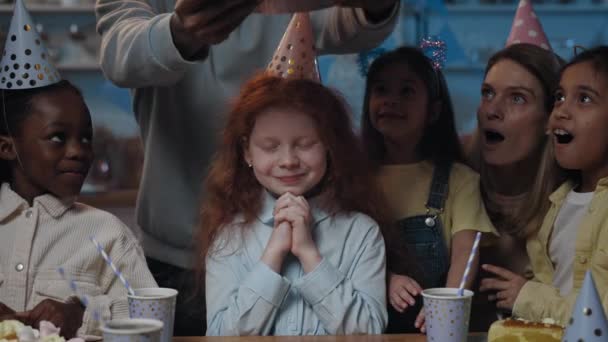 Vista de la cosecha de lindo niño pequeño con el pelo rojo esperando con los ojos cerrados, mientras que el hombre trae pastel de cumpleaños en frente de ella. Chica soplando velas en la torta y durante la fiesta. — Vídeos de Stock