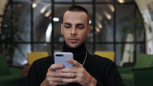 Vista de la cosecha de tipo milenario con cerdas usando el teléfono inteligente y sonriendo mientras está sentado en la cafetería. Hombre guapo con una cadena escribiendo un mensaje mientras charlaba en las redes sociales. Concepto de ocio. — Vídeo de stock