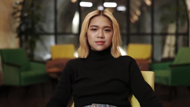 Crop view of young woman with blond dyed hair and make up looking to camera. Portrait of pretty female person posing while sitting in chair at cafe. — Stock Video