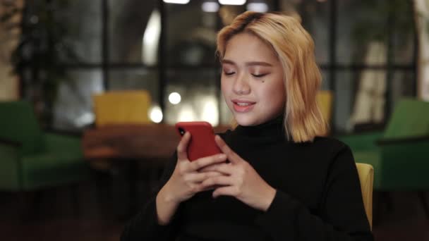 Vista de cerca de la mujer milenaria con el pelo corto teñido rubio usando el teléfono inteligente mientras está sentado en la silla. Persona bastante femenina sonriendo mientras chatea en las redes sociales en la cafetería. — Vídeos de Stock