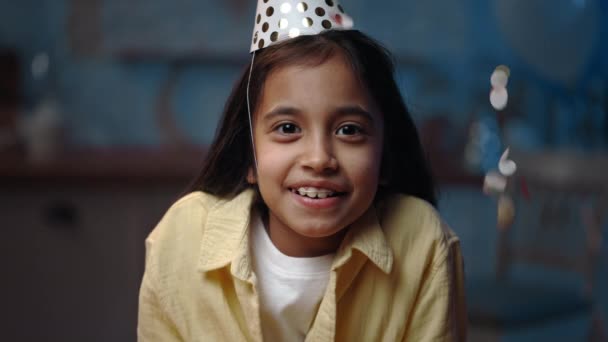 Vista de la cosecha de niña positiva sonriendo y mirando a la cámara. Lindo niño femenino en el sombrero de cumpleaños regocijo y posando mientras confeti caer fondo borroso. — Vídeo de stock