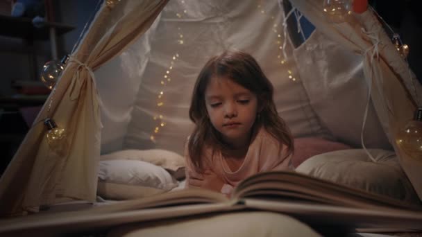 Hübsches kleines Mädchen in niedlichen Zeit in dekorativen provisorischen Hütte zu Hause am Abend verbringen. Comely Kind auf dem Boden liegend beim Lesen von Büchern. Konzept der Freizeit und der sorglosen Kindheit. — Stockvideo