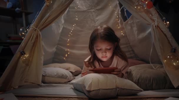 Linda niña pasando tiempo en la cabaña improvisada decorativa en casa por la noche. Niño atractivo tumbado en el suelo mientras ve dibujos animados en smartphone.Concept de ocio y la infancia descuidada. — Vídeo de stock