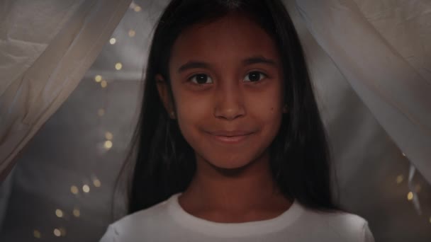 Retrato de menina adolescente bonito com cabelo preto longo olhando para a câmera. Headshot de adolescente muito feminina em tenda decorativa com iluminações.. — Vídeo de Stock