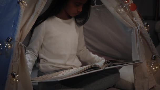 Menina bonita na cabana improvisada decorativa lendo livro em casa à noite. Adolescente feminina com longos cabelos pretos sentados no chão enquanto lê o livro. Conceito de lazer. — Vídeo de Stock