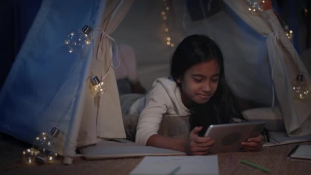 Menina adorável em tenda decorativa bonito assistindo desenhos animados em casa à noite. Muito adolescente deitado no chão ao usar tablet. Conceito de lazer e infância descuidada. — Vídeo de Stock