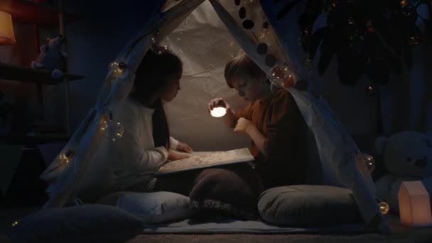 Adolescentes em cabana improvisada decorativa olhando para a página do livro à noite. Menino e menina usando lanterna enquanto sentado no chão e passar o tempo livre juntos. Conceito de lazer. — Vídeo de Stock