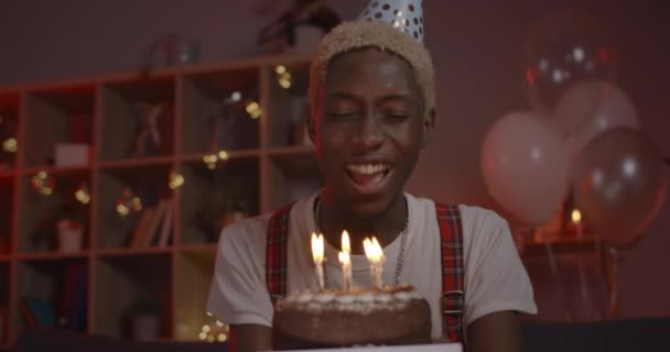 Крутий погляд на веселого афро-американського чоловіка в капелюсі до дня народження радіючи і розмовляючи, дивлячись на камеру. Молодий приємний хлопець з пофарбованим волоссям, який віє свічки на торті, сидячи вдома.. — стокове відео
