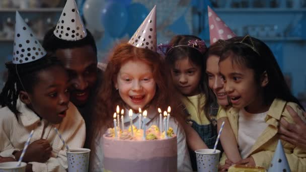 Вид на різноманітних дітей, чоловік і жінка дме свічки на торт разом з дівчиною на день народження. Щасливі люди та діти у святкових шапках проводять вечірку. Концепція святкування . — стокове відео