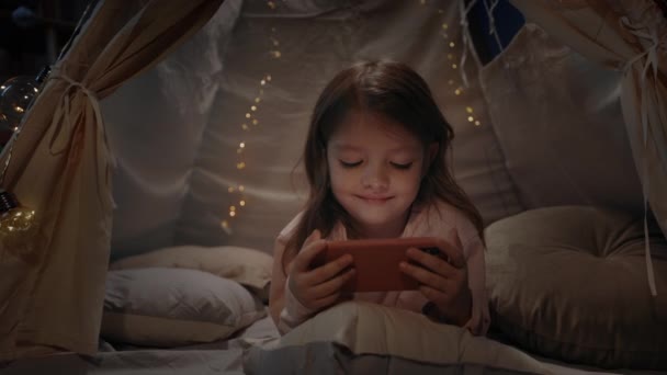 Vrolijk klein meisje brengt tijd door in decoratieve tent met verlichting in de avond speelkamer. Kind liggend op de vloer en glimlachend tijdens het kijken naar cartoon op smartphone. Het begrip kindertijd. — Stockvideo