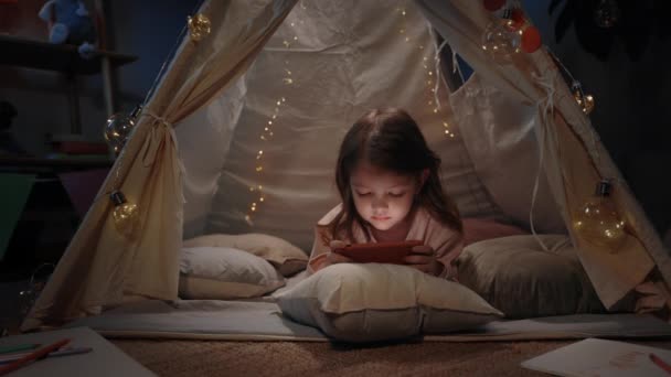 Schattig meisje in mooie geïmproviseerde hut spelen spel online thuis in de avond. Mooi klein kind liggend op de vloer tijdens het gebruik van de smartphone. Concept van vrije tijd en zorgeloze jeugd. — Stockvideo