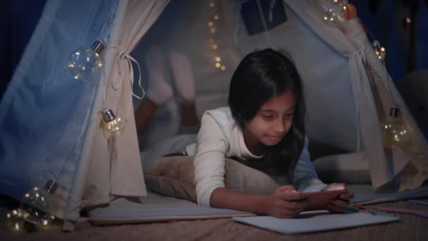 Fille gaie dans une cabane de fortune décorative avec éclairage regarder la vidéo à la maison le soir. Jolie adolescente allongée sur le sol tout en utilisant l'écran du téléphone Concept de loisirs. — Video