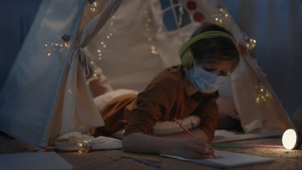 Підліток з медичною захисною маскою малює кольоровими олівцями, лежачи на підлозі в декоративному наметі. Хлопчик-підліток слухає музику в навушниках, проводячи вільний час . — стокове відео