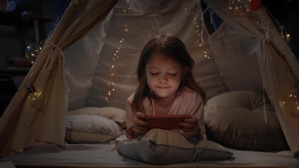 Schattig meisje in leuke decoratieve tent spelen online games thuis in de avond. Klein kind liggend op de vloer tijdens het gebruik van smartphone. Concept van vrije tijd en zorgeloze jeugd. — Stockvideo