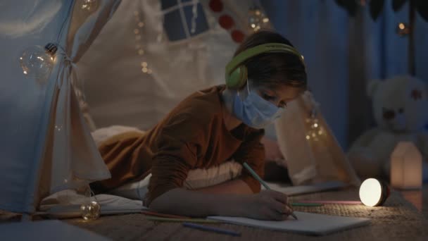 Ung pojke med medicinsk skyddsmask ritning med färgade pennor när du ligger på golvet i dekorativa tält. Smart manlig tonåring lyssnar på musik i hörlurar medan du tillbringar fritid. — Stockvideo