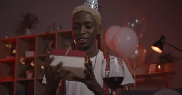 Felice uomo afro americano in cappello di compleanno guardando alla fotocamera e sorridente. Persona maschile gioiosa che parla e scuote la scatola regalo mentre fa conversazione online a casa. — Video Stock