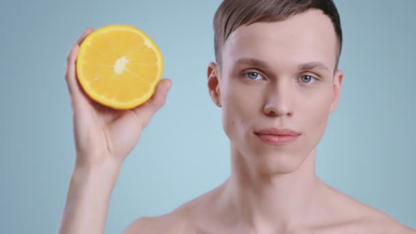 长相英俊的男人一边拿着一半橙子一边看着相机的画像。男模，光着肩膀，淡蓝色的眼睛，摆设着水果。护肤、保健的概念. — 图库视频影像