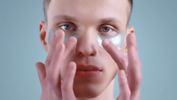 Snímek hlavy mladého muže, který se dotýká hydropeptidových skvrn pod očima, zatímco se dívá do kamery. Portrét pohledného mužského modelu se světle modrýma očima pózujícího. Koncepce péče o pleť. — Stock video