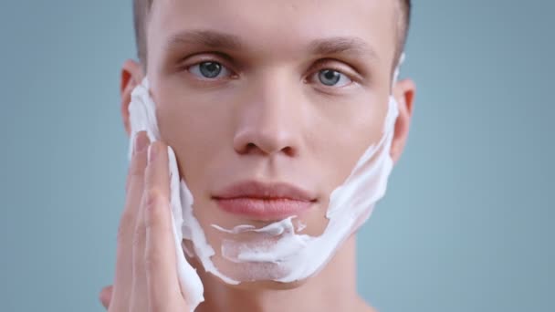 Tiro na cabeça de um jovem olhando para a câmera enquanto coloca espuma branca de limpeza ou barbear no rosto. Retrato de modelo bonito masculino com olhos azuis claros posando. — Vídeo de Stock