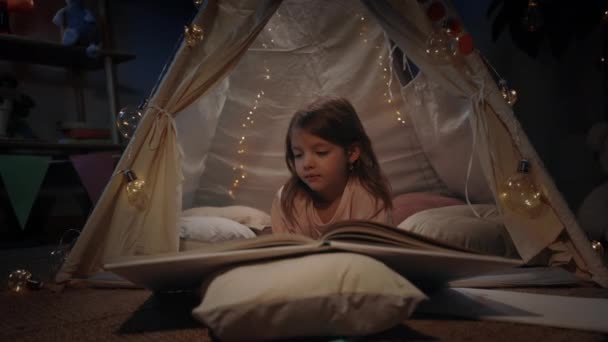 Aranyos lány gyönyörű makeshift kunyhó fordult oldalak otthon este. Csinos kisgyerek fekszik a földön, miközben könyvet olvas. A szabadidő és a gondatlan gyermekkor fogalma. — Stock videók