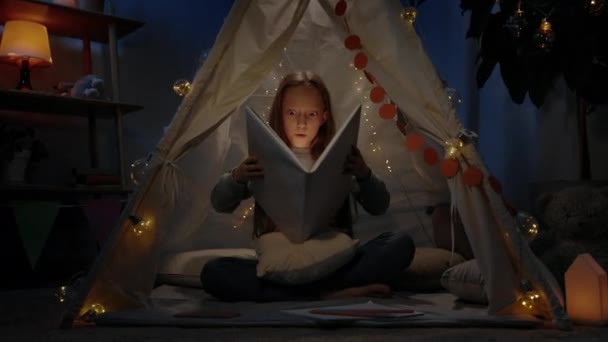 Niesamowita dziewczyna z czerwonymi włosami w prowizorycznym namiocie otwierającym książkę w domu wieczorem. Nastolatek robi zaskoczoną minę siedząc na podłodze i czytając książkę z latarką. — Wideo stockowe