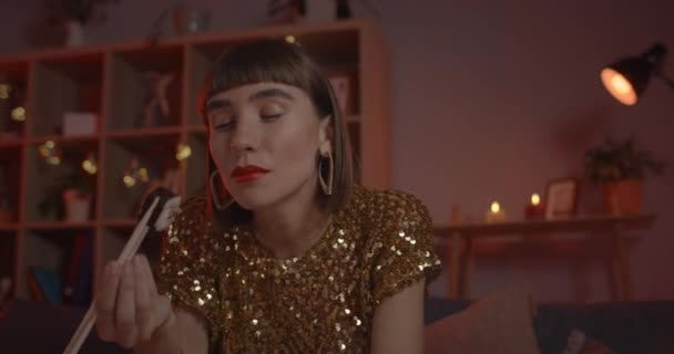 Vista de la cosecha de la mujer joven comiendo rollos de sushi mientras mantiene una conversación en línea. Persona femenina con ropa elegante sonriendo y hablando mientras mira a la cámara en casa. Concepto de fecha en línea. — Vídeos de Stock