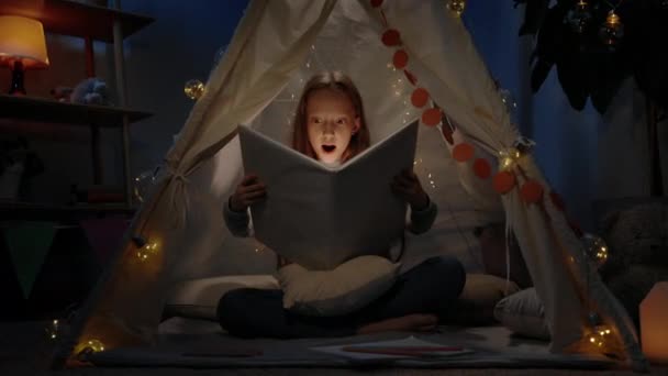 Ładna dziewczyna z czerwonymi włosami w prowizorycznym namiocie otwierającym książkę w domu wieczorem. Ciekawy nastolatek robi zaskoczoną minę siedząc na podłodze i czytając książkę z latarką. — Wideo stockowe