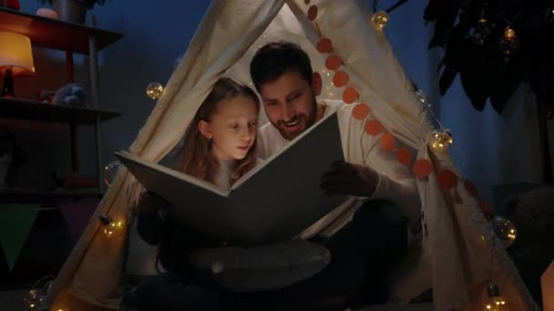 Schöner Vater und sein Teenie-Mädchen öffnen gemeinsam ein Buch, während sie in einem dekorativen Behelfszelt sitzen. Eine glückliche Familie, die Märchen liest und gleichzeitig eine schöne Zeit miteinander verbringt. Konzept reales Leben. — Stockvideo