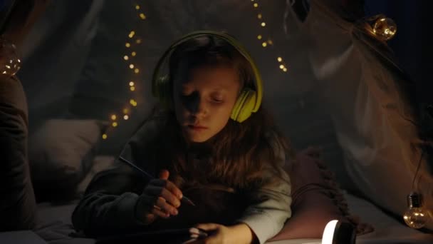 Όμορφο κορίτσι με ακουστικά που σκέφτεται και γράφει ενώ βρίσκεται στο πάτωμα σε διακοσμητική αυτοσχέδια σκηνή στο σπίτι. Έφηβοι ακούνε μουσική ενώ περνούν ελεύθερο χρόνο το βράδυ. Έννοια του ελεύθερου χρόνου. — Αρχείο Βίντεο