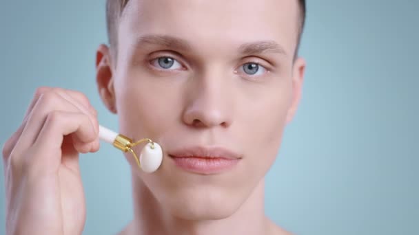 얼굴 마사지 롤러를 사용하는 젊은 남자의 초상화 카메라를 찾는 동안. 밝은 파란색 눈 포즈를 한 남자 밀레니엄 모델 .Concept of skincare, face care. — 비디오
