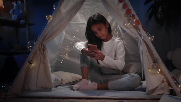 一个长黑头发的漂亮女孩晚上呆在家里装饰临时搭建的小木屋里。青少年坐在地板上观看智能手机的使用和滚动。休闲的概念. — 图库视频影像
