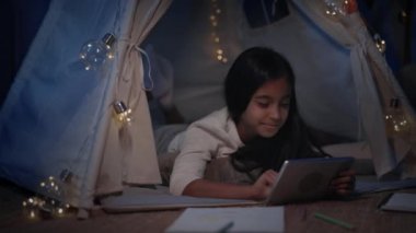 Neşeli kız akşamları evdeki dekoratif derme çatma kulübede vakit geçiriyor. Tablette video izlerken yerde yatan güzel bir genç. Boş zaman ve dikkatsizlik kavramı.