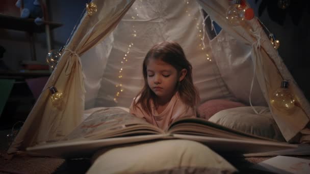 Menina adorável na tenda decorativa bonito virando páginas em casa à noite. Criança deitada no chão enquanto lia um livro interessante. Conceito de lazer e infância descuidada. — Vídeo de Stock