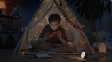Akşamları dekoratif çadırda oyun oynayan sarı saçlı heyecanlı genç çocuk. Akıllı telefon kullanırken tennager yerde oturuyor. Boş zaman ve boş zaman kavramı..