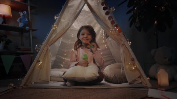 Adorable niña en hermosa carpa decorativa que sopla bubbless y mirando a la cámara en la noche. Niña sentada en el suelo mientras se divierte en su habitación. Concepto de infancia. — Vídeo de stock