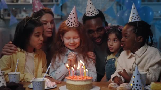 Criança bonita com longos cabelos vermelhos esperando com os olhos fechados, enquanto o homem trazendo bolo de aniversário na frente dela. Menina soprando velas no bolo e durante a festa. — Vídeo de Stock