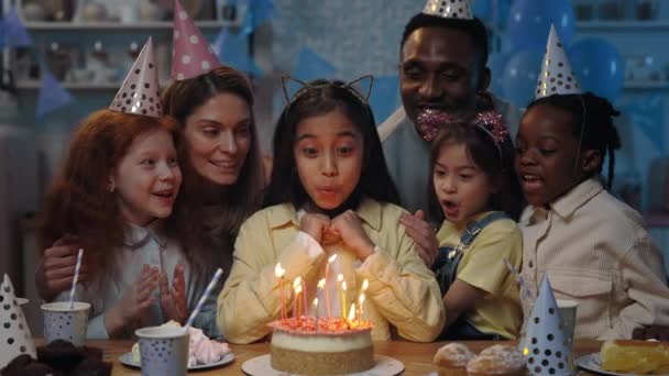 Вид на коріння жінки, яка робить бажання і дме свічки на торт разом з багатьма етнічними дітьми і дорослими. Чоловік, жінка і діти зі святковими шапками, що проводять вечірку на день народження . — стокове відео