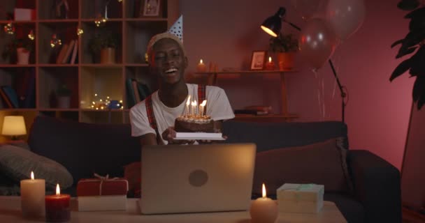 Bonne personne masculine afro-américaine avec gâteau d'anniversaire dans les mains accepte les salutations par Webcam. Un homme célèbre virtuellement avec sa famille en utilisant un ordinateur portable en quarantaine, éteint des bougies, fait un vœu. — Video