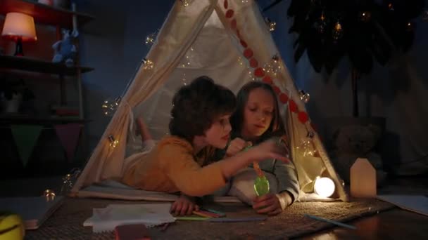 Dzieci w cute dekoracyjne prowizoryczne chata dmuchanie bąbelków w domu wieczorem. Dziewczyna i chłopiec leżą na podłodze i dobrze się bawią. Pojęcie czasu wolnego i niedbałego dzieciństwa. — Wideo stockowe
