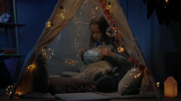 Gadis remaja mengenakan topeng medis untuk boneka beruang sambil duduk di lantai di tenda darurat dekoratif di rumah. Anak dengan mainan stetoskop bermain di malam hari. Konsep waktu luang. — Stok Video