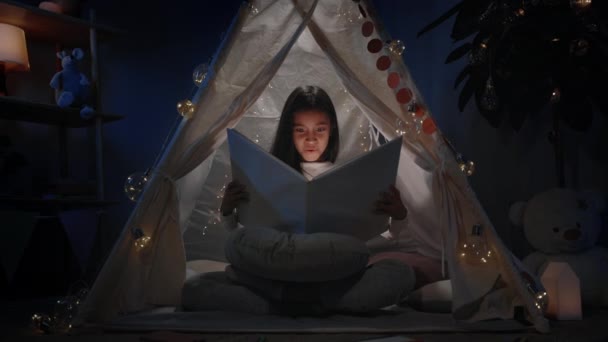 Ragazza eccitata con lunghi capelli neri in simpatico rifugio libro apertura a casa in serata. Carino grazioso adolescente seduto sul pavimento durante la lettura di un libro con torcia elettrica. Concetto di tempo libero e tempo libero. — Video Stock