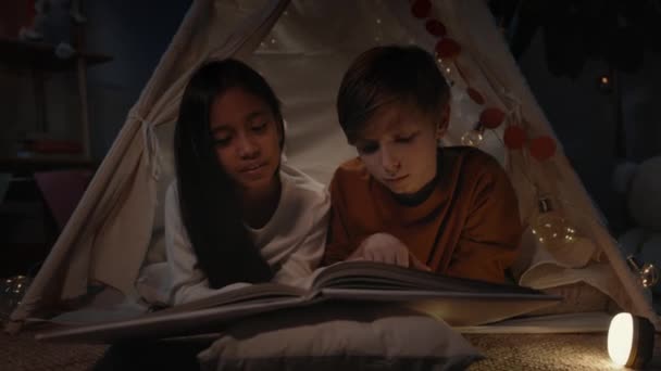 Adolescentes en cabaña improvisada decorativa leyendo libro y sonriendo en casa por la noche. Niño y chica positivos tumbados en el suelo mientras pasan tiempo libre. Concepto de ocio y descuido. — Vídeo de stock