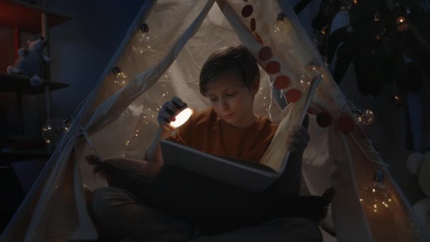 Mladý chytrý chlapec s blond vlasy v dekorativním stanu čtení zajímavé knihy doma ve večerních hodinách. Teenager sedí na podlaze a tráví volný čas. Pojetí volného času. — Stock video