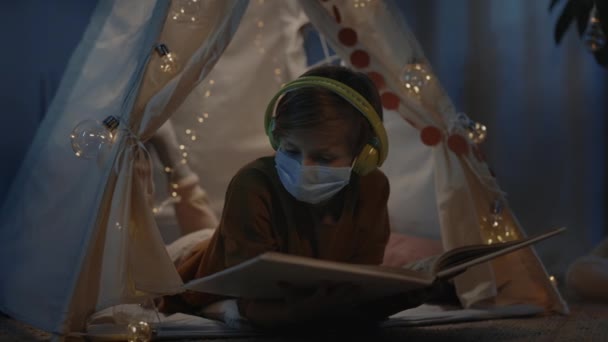 Niño con auriculares pasar tiempo en la tienda improvisada decorativa en casa por la noche. Adolescente con máscara médica acostada en el suelo mientras lee un libro. Concepto de ocio. — Vídeos de Stock